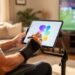 Person wearing a wrist brace uses a cushioned stylus on a tablet mounted on an adjustable stand at eye level, seated comfortably in a living room with a blurred sports broadcast on the TV in the background.